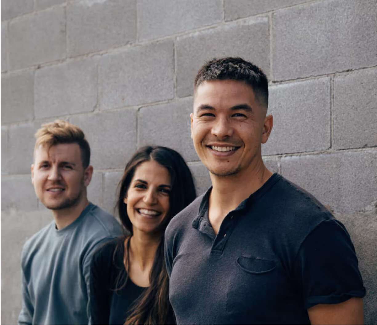 Three people smiling and standing against a gray brick wall, looking at the camera.