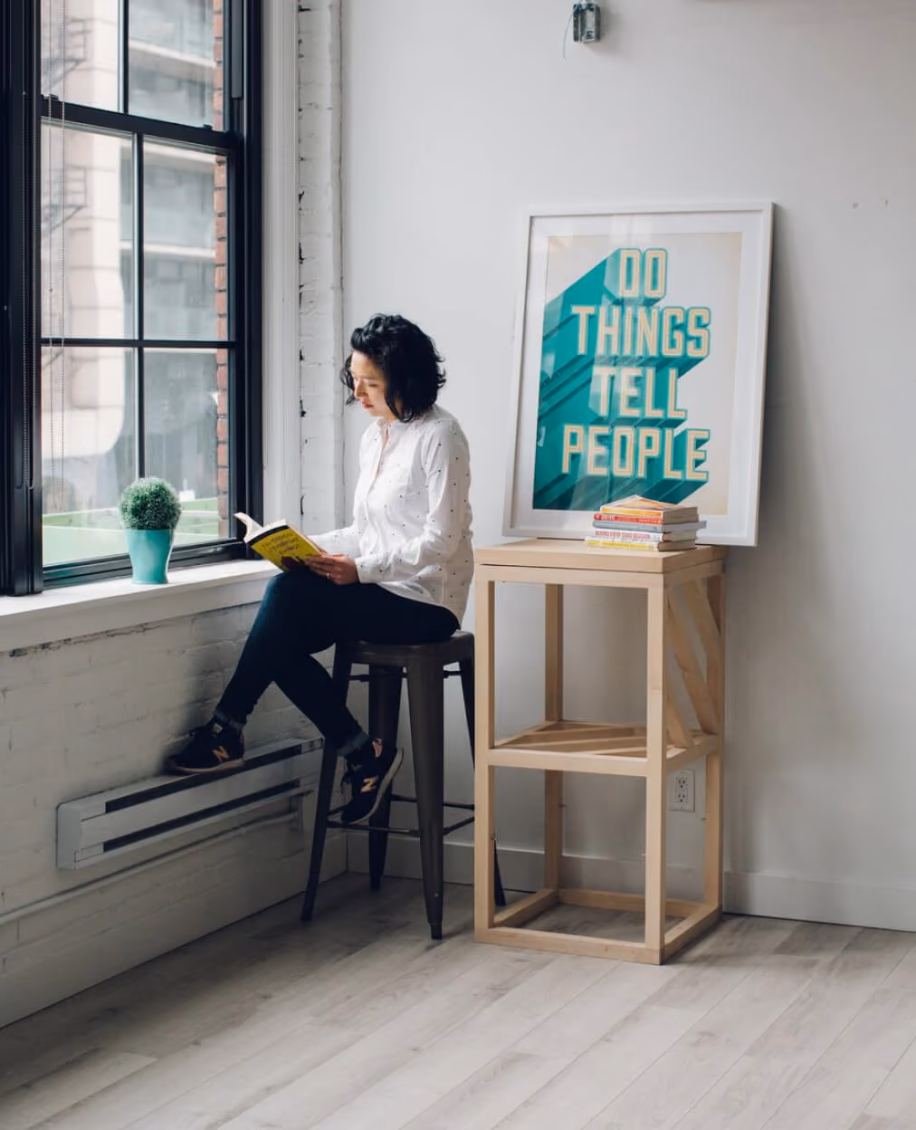 Woman sitting on stool by a large window reading a book next to a wooden table with a framed print.