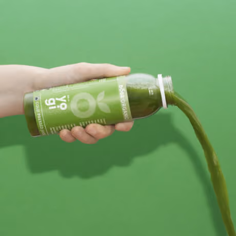 Hand pouring green organic cold-pressed juice from a bottle against a green background.