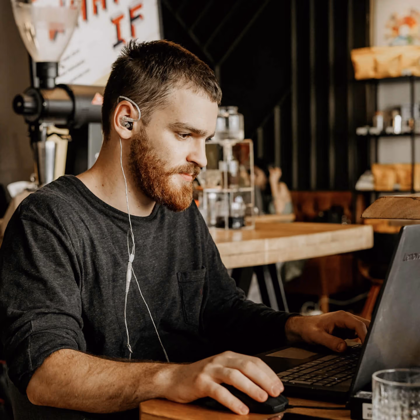 Man with a beard wearing earphones, working on a laptop in a cozy indoor café.