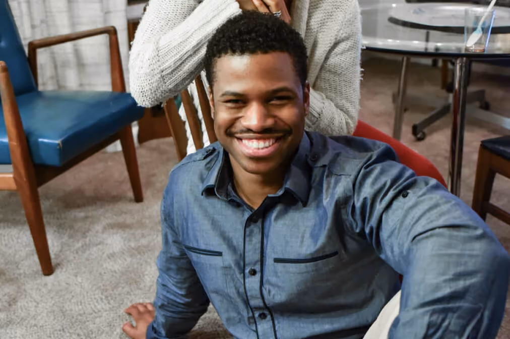 Smiling man sitting on carpeted floor with person behind wearing white sweater.