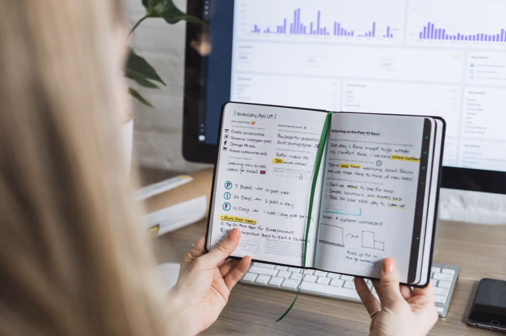 Person holding open notebook with handwritten notes in front of monitor showing charts on desk.