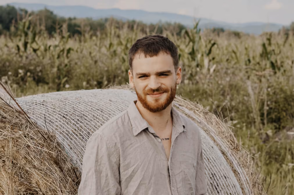 Smiling young man with beard standing outdoors near wrapped hay bales with trees in the background.