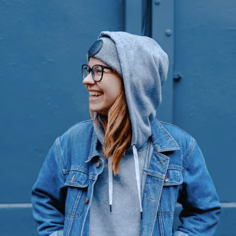 Smiling woman wearing glasses, a gray beanie, hoodie, and denim jacket against a blue background.