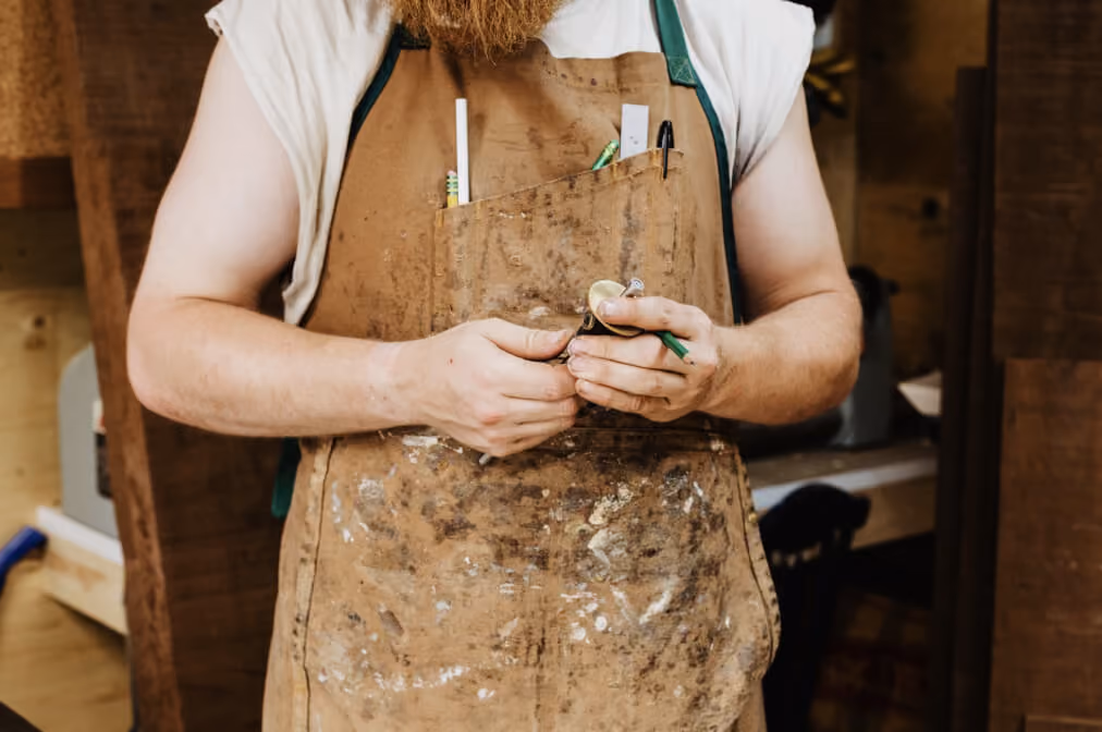 Bearded man in a stained brown apron holding a small metal object inside a wooden workshop.