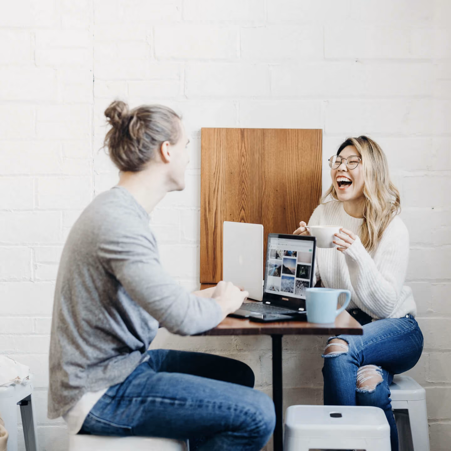 Two people at a table with laptops, one woman laughing holding a coffee cup and a man facing her.