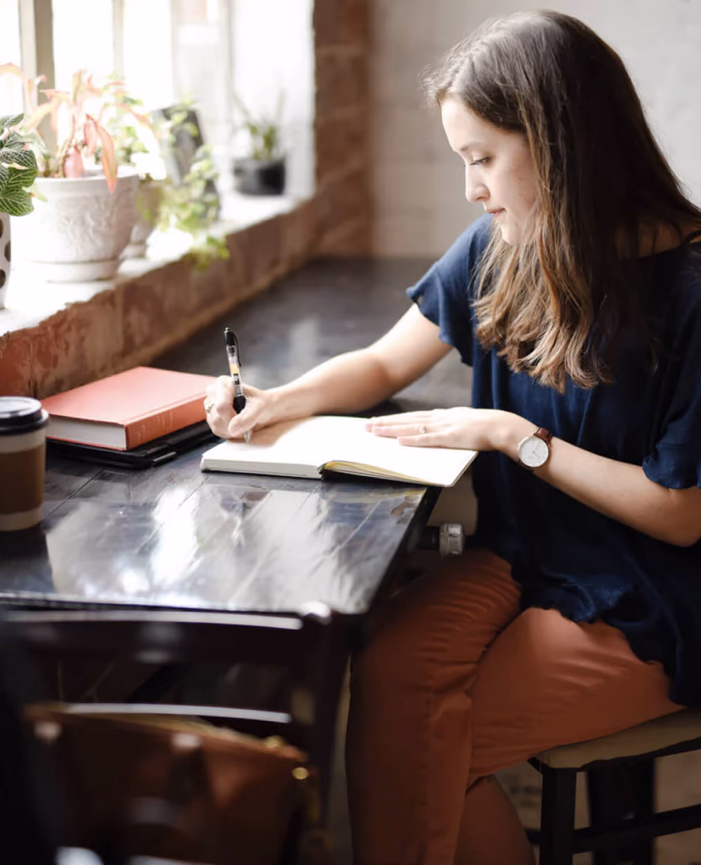 Young woman writing in a notebook at a wooden table near a window with potted plants.
