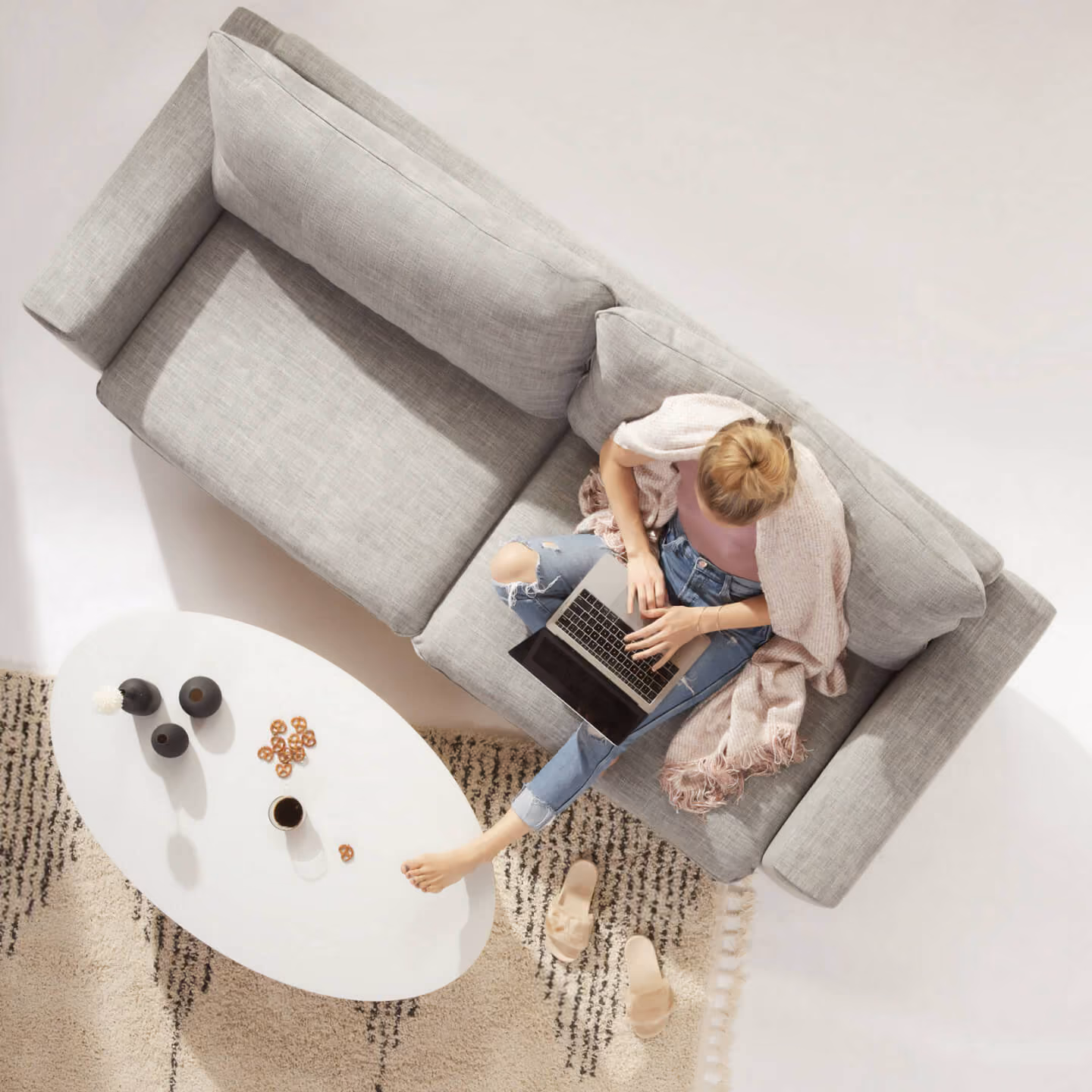 Woman sitting on gray couch with laptop, feet on rug near white table with coffee and snacks.