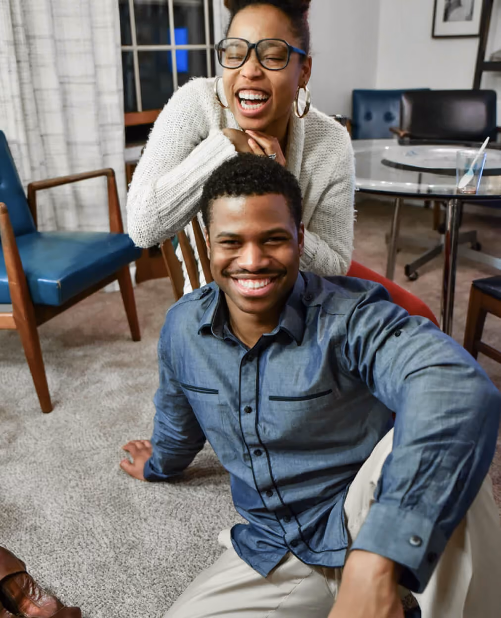 Smiling man sitting on carpet with woman behind laughing joyfully in a cozy room.