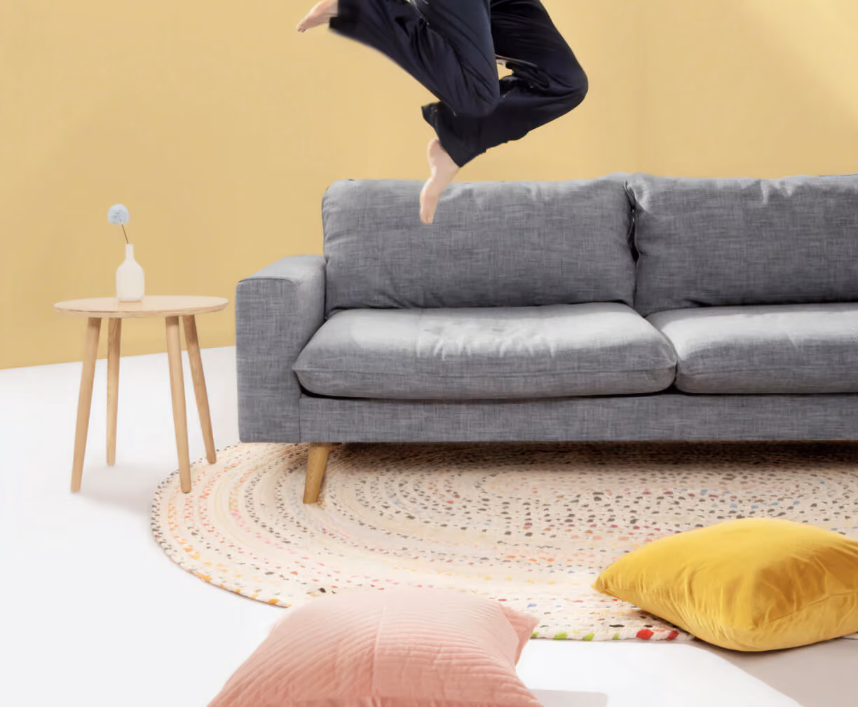 Person jumping barefoot above gray sofa in a bright room with round rug and pillows on floor.