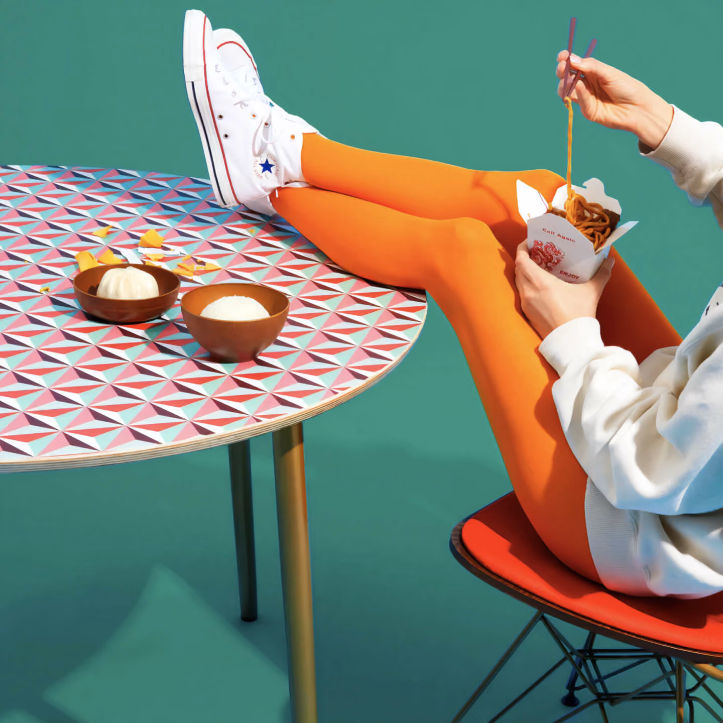 Person in orange tights and white sneakers eating noodles with chopsticks, legs on patterned table.