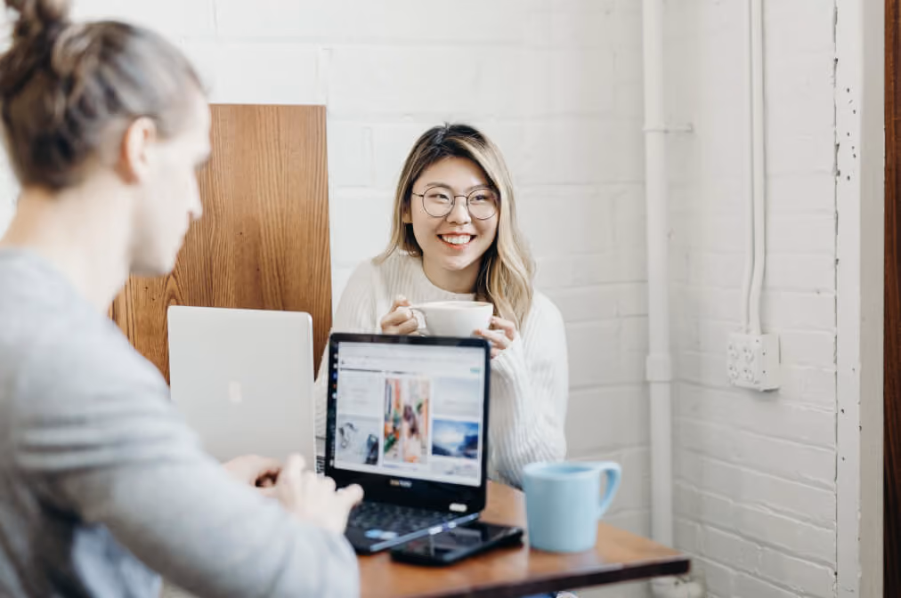 Two people working with laptops in a bright coffee shop, one smiling while holding a cup.