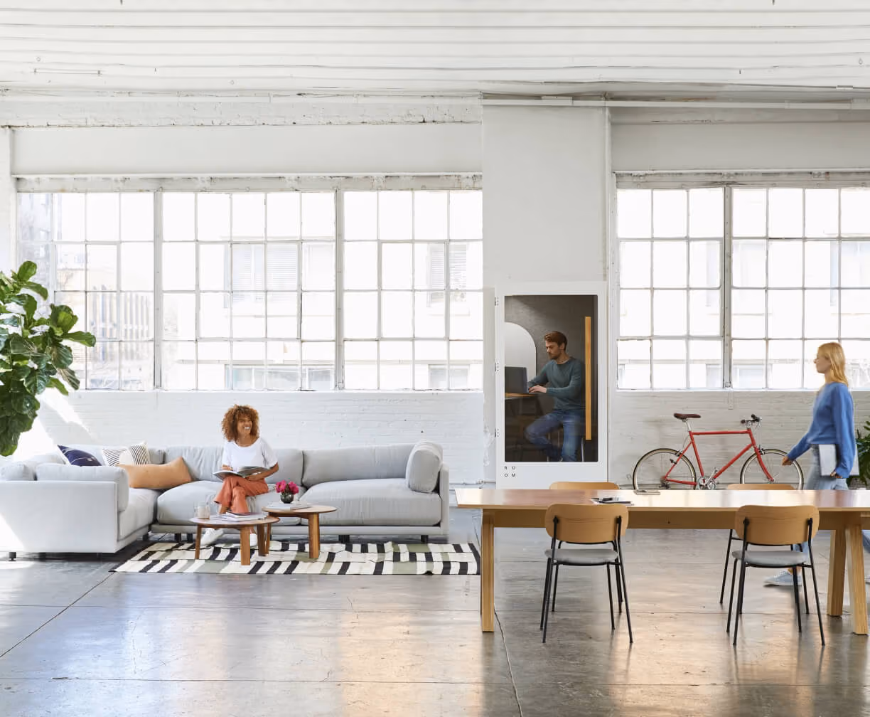 Modern office with woman sitting on a couch, man working in phone booth, and woman walking by a red bicycle.