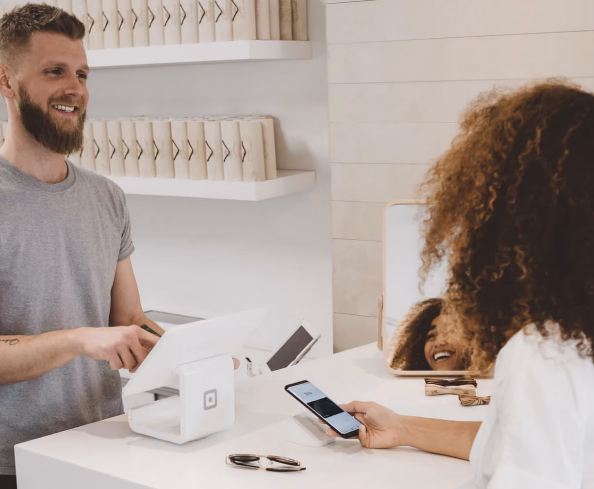Smiling man at a counter assists woman with curly hair paying using smartphone contactless method.