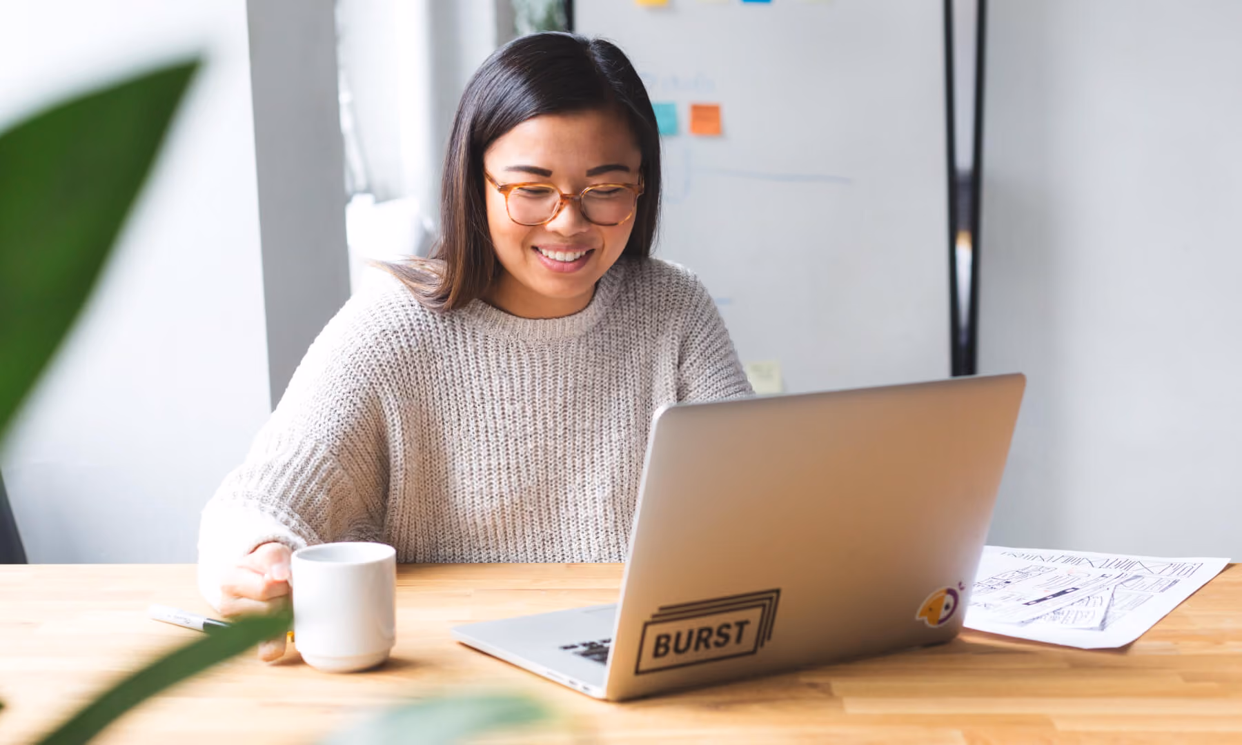 Smiling woman wearing glasses, working on a laptop at a wooden table with a mug in hand.