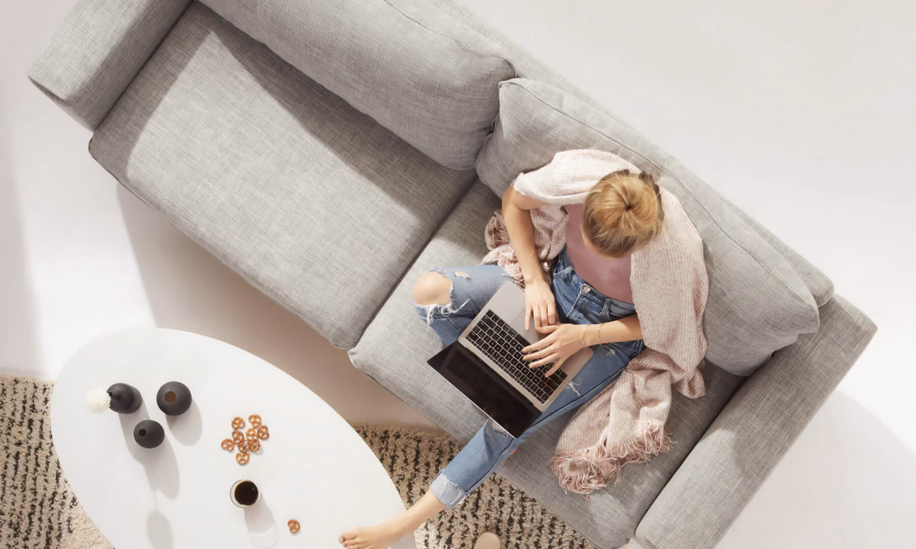Top view of a woman with blonde hair in a bun sitting on a gray couch working on a laptop.
