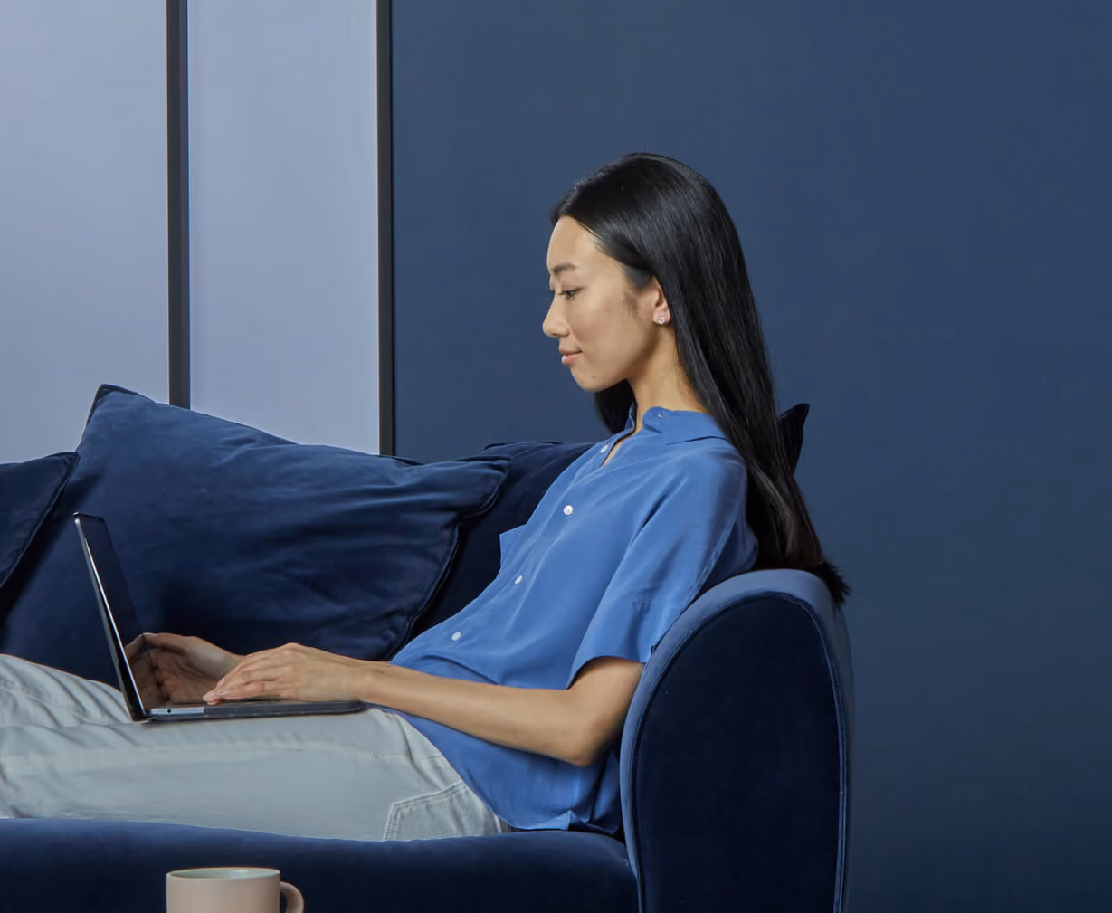 Woman in blue shirt sitting on navy sofa using a laptop with a mug in the foreground.