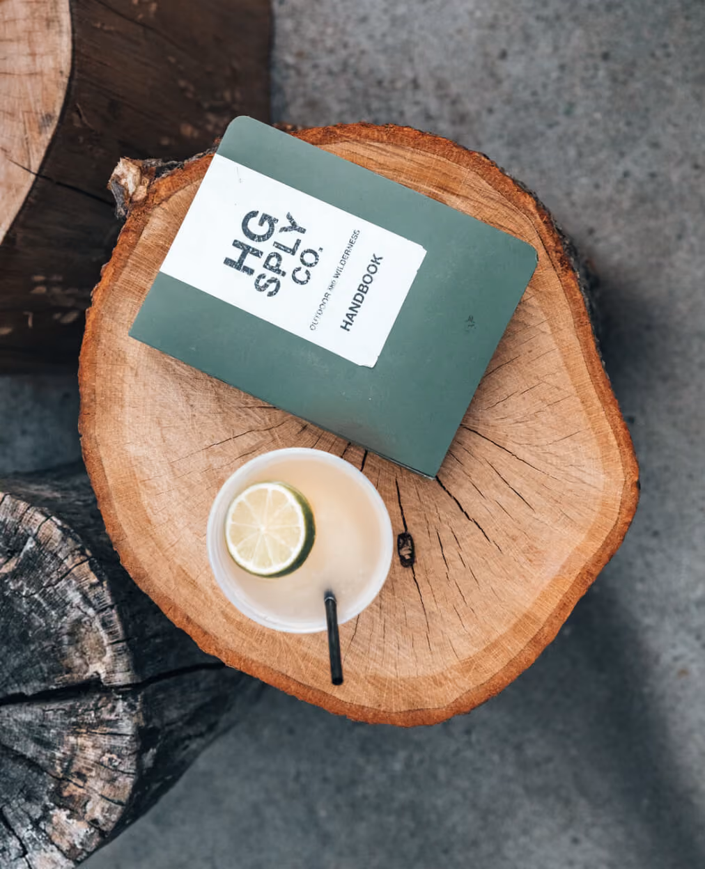Green handbook and cup with a lime slice and black straw on a round wooden log table.