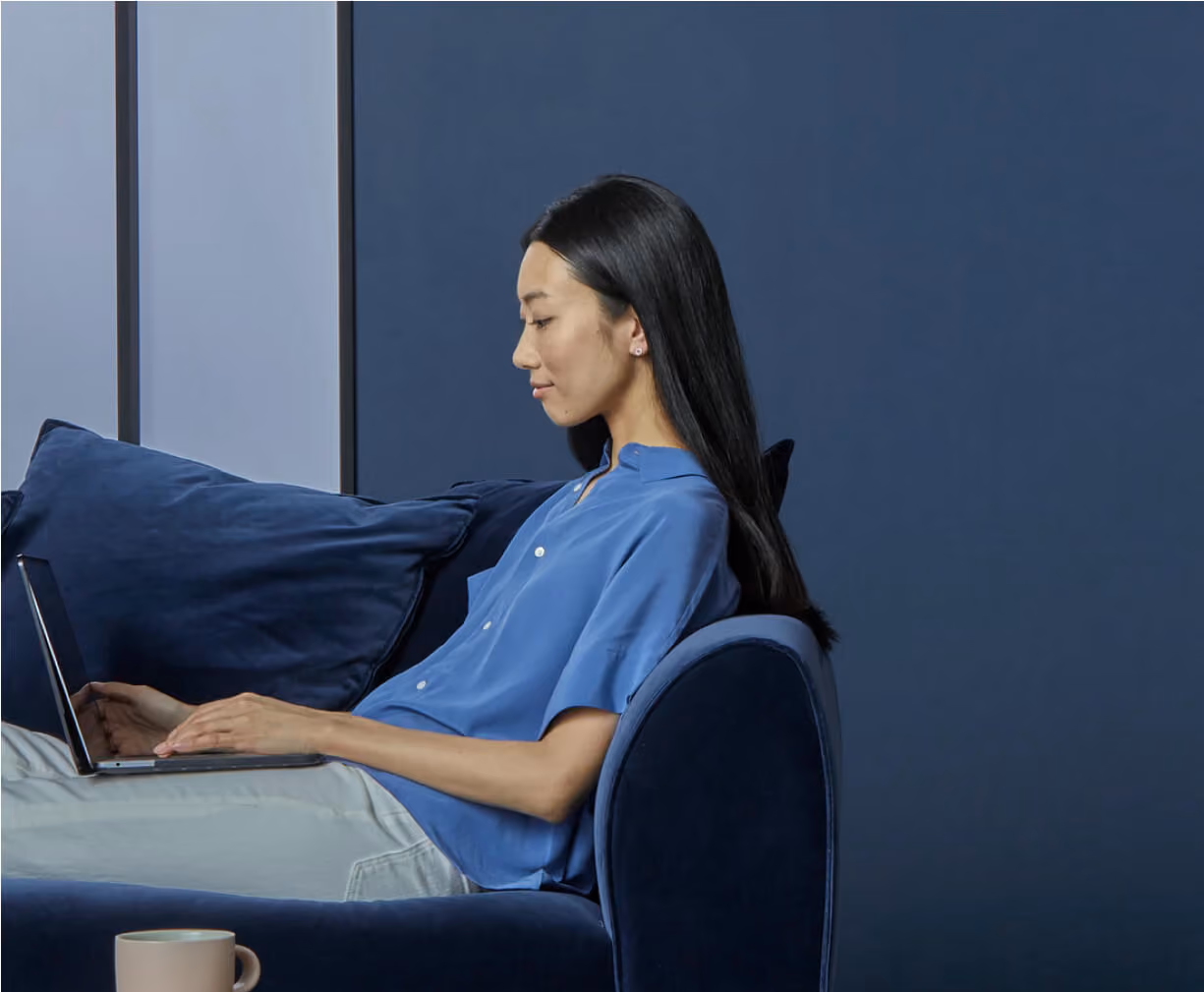Woman in blue shirt sitting on navy sofa using a laptop with a beige mug on the armrest.
