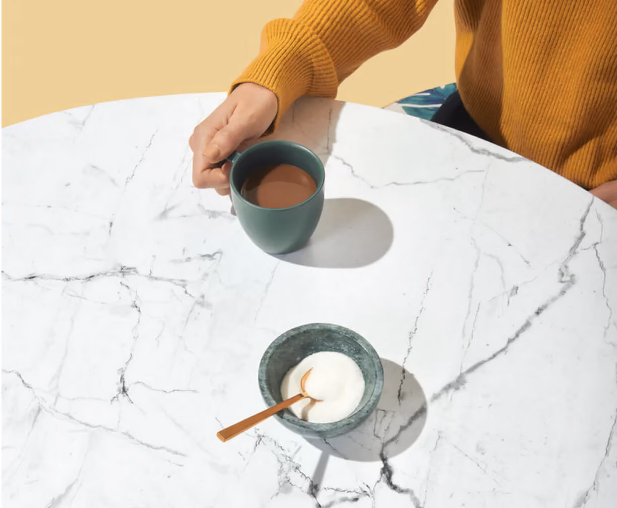 Person in mustard sweater holding a green mug of coffee on a white marble table with a bowl of sugar.