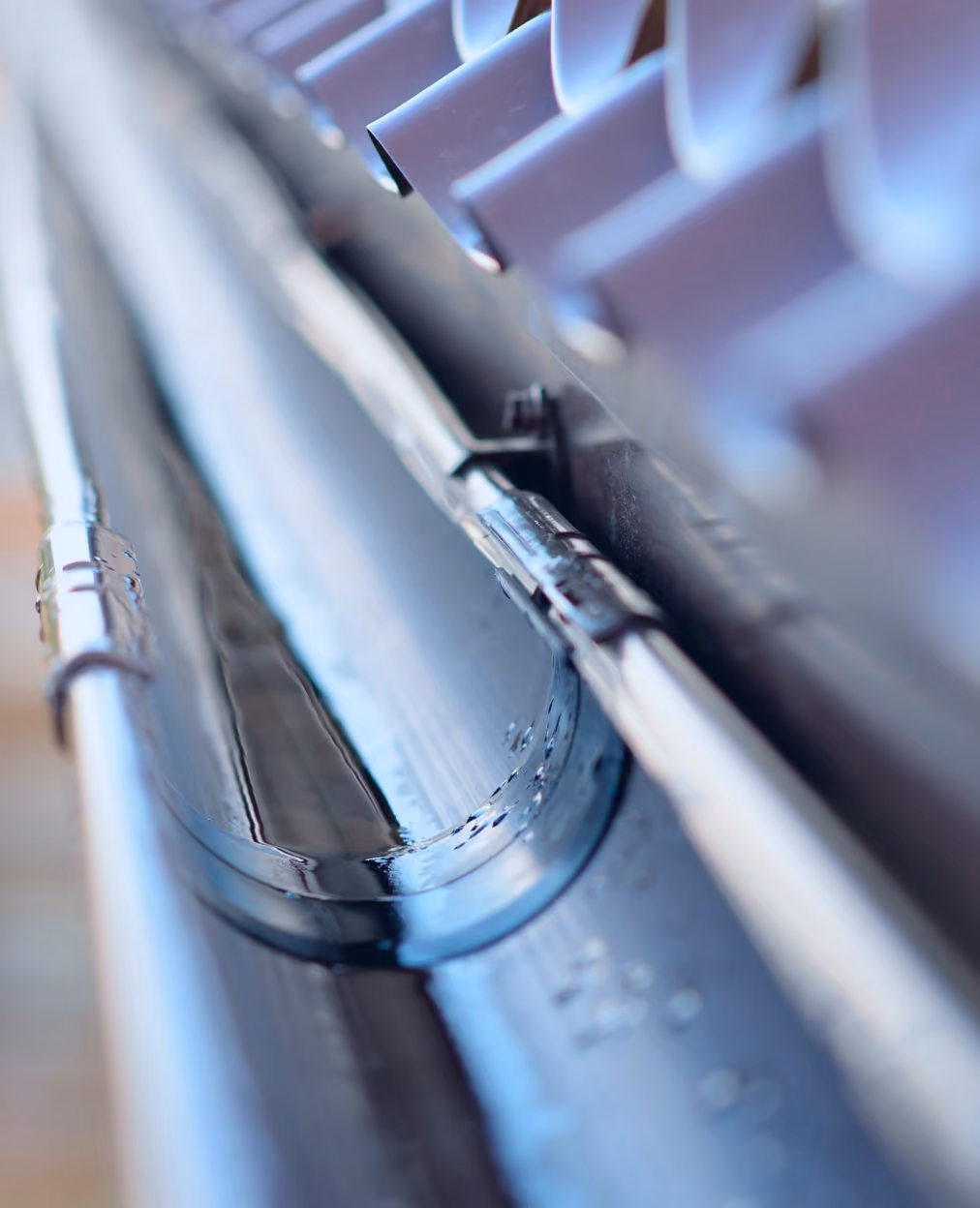 Close-up of a clean, wet metal roof gutter with attached piping and roof tiles above.