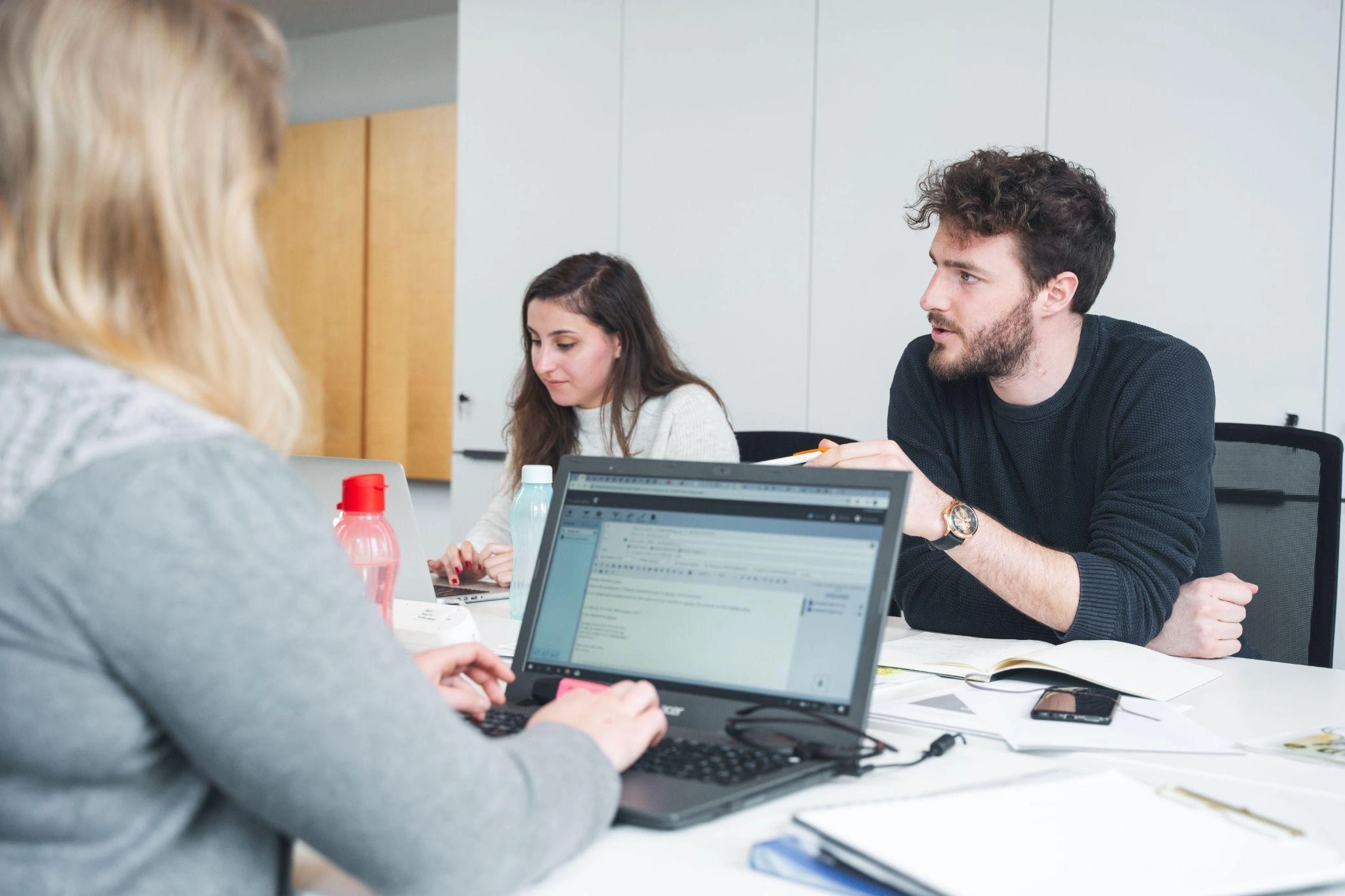 Trois jeunes adultes travaillant ensemble autour d'une table avec des ordinateurs portables, des carnets et des bouteilles d'eau dans un bureau moderne.