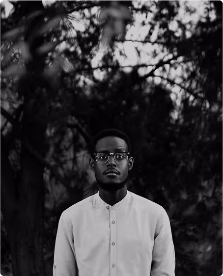 Man with glasses and a buttoned-up shirt standing outdoors with trees in the background, in black and white.