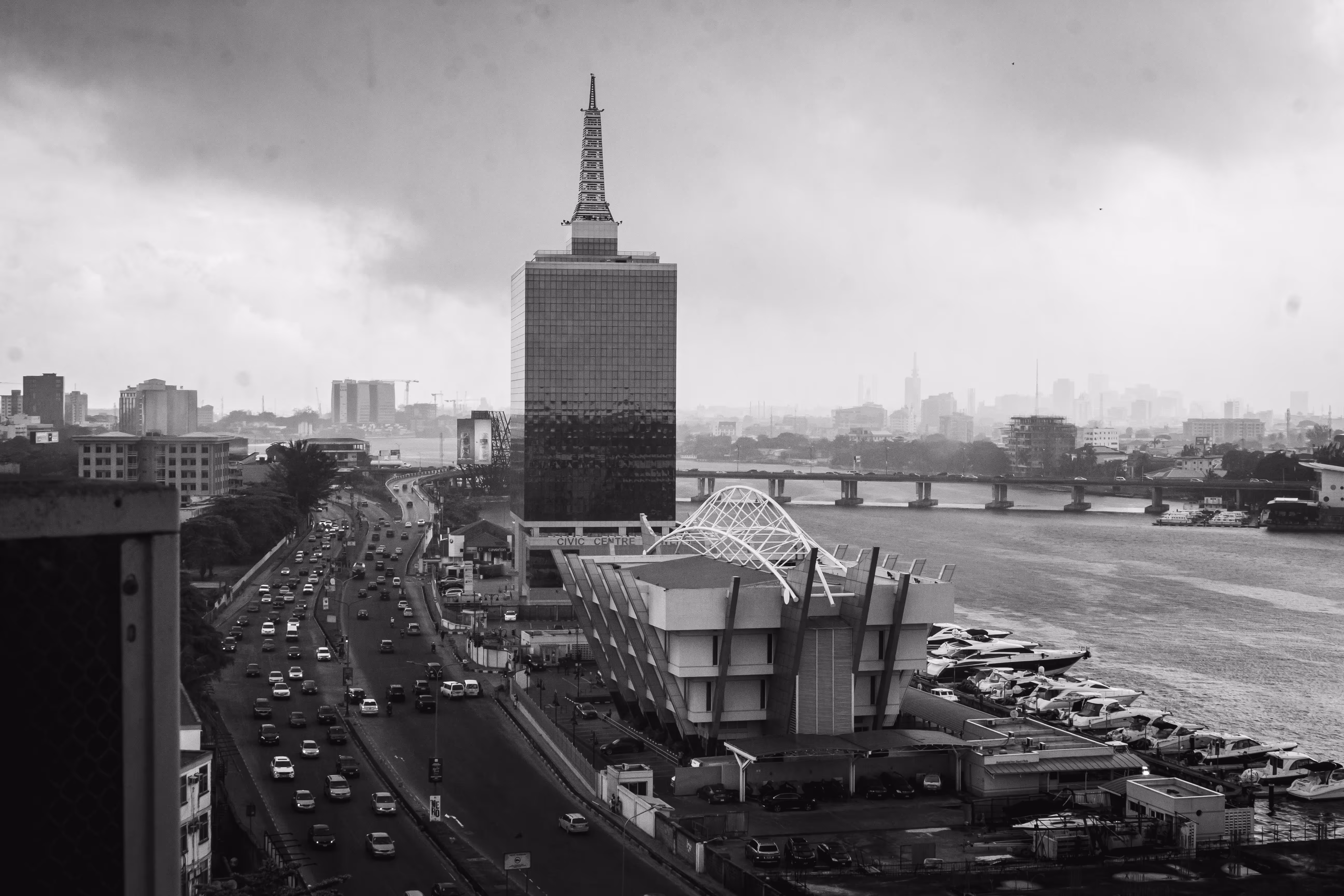 Black and white skyline view of Lagos with a tall building, busy road, modern architecture, and a river with docked boats.