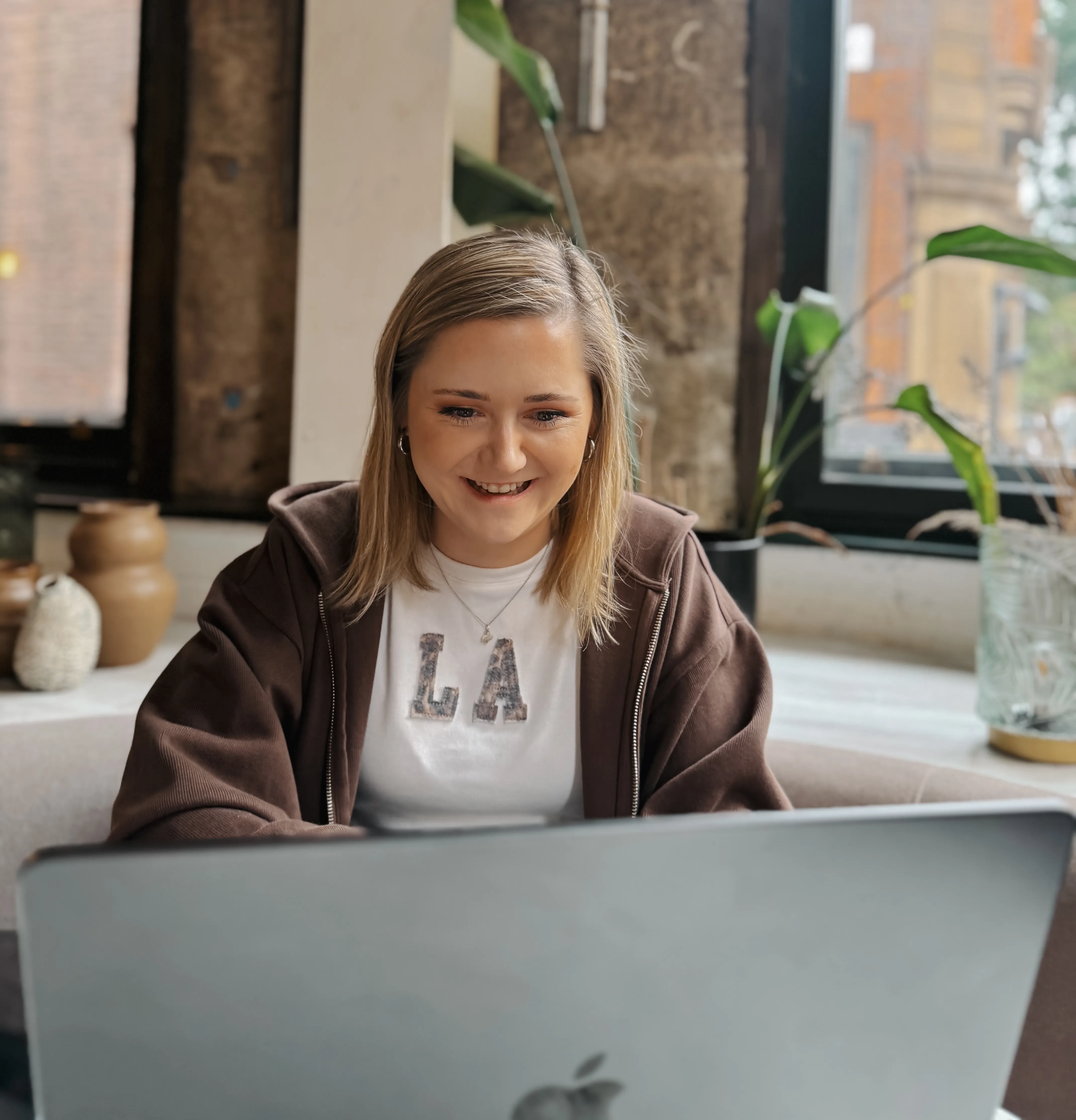 Smiling woman working on a laptop indoors near a window.