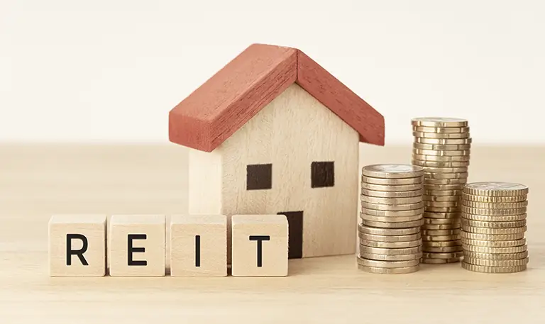 Къща и монети до буквите REIT, символизиращи тръст за недвижими имоти и доходи от наем.