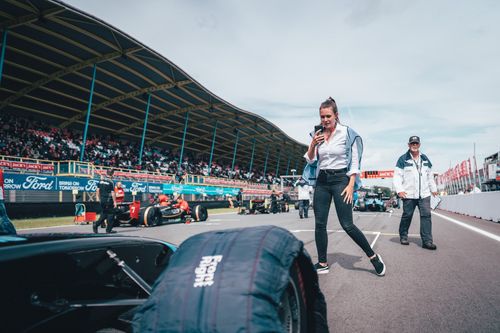 Woman on racetrack taking a video on her phone during motorsport event, with cars and crew in the background.