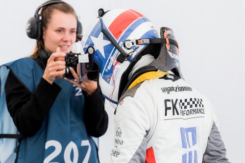 A racedriver being filmed by a woman with a camera in her hand.