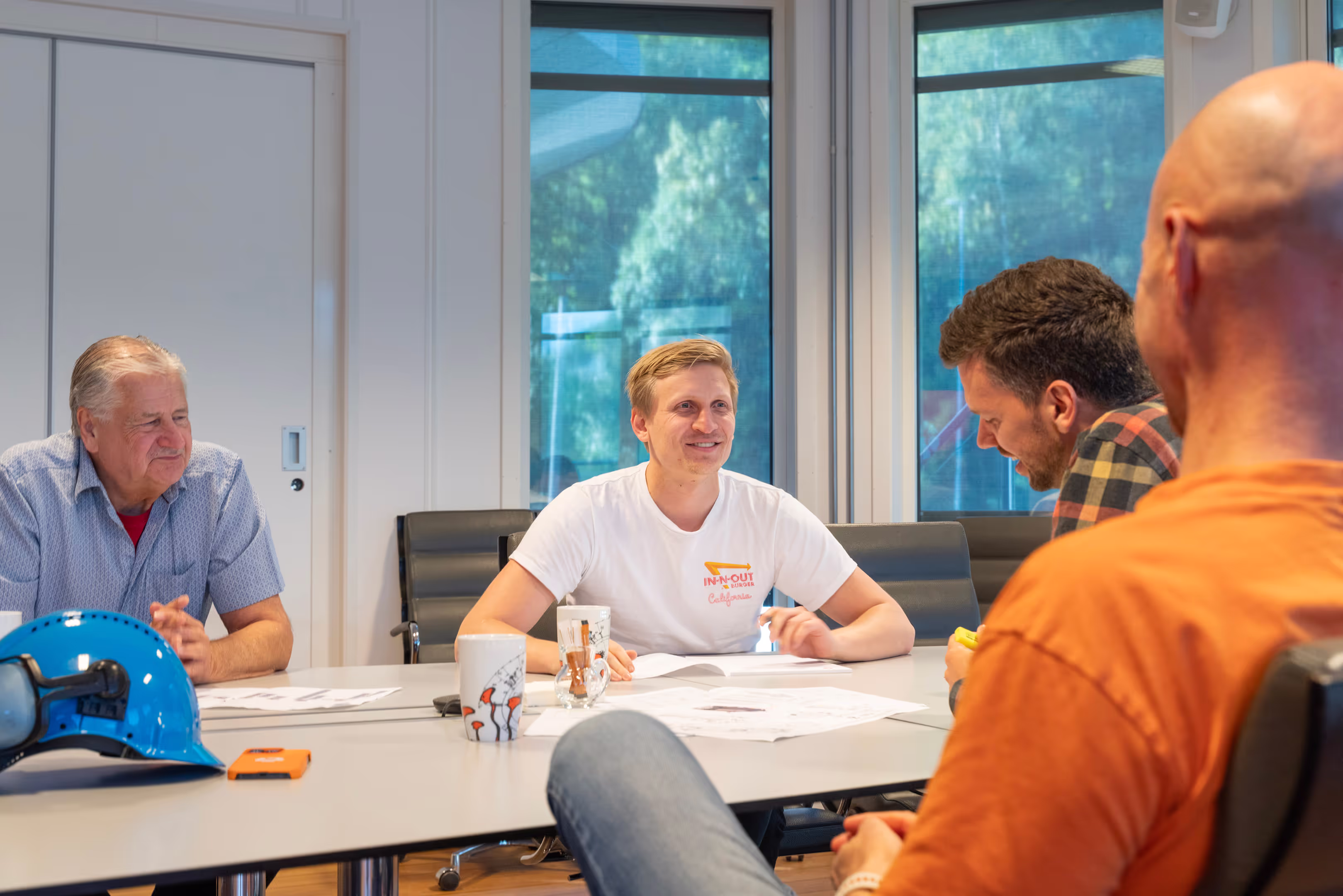 Four men sitting around a conference table in a meeting room with papers, mugs, and a blue safety helmet on the table.