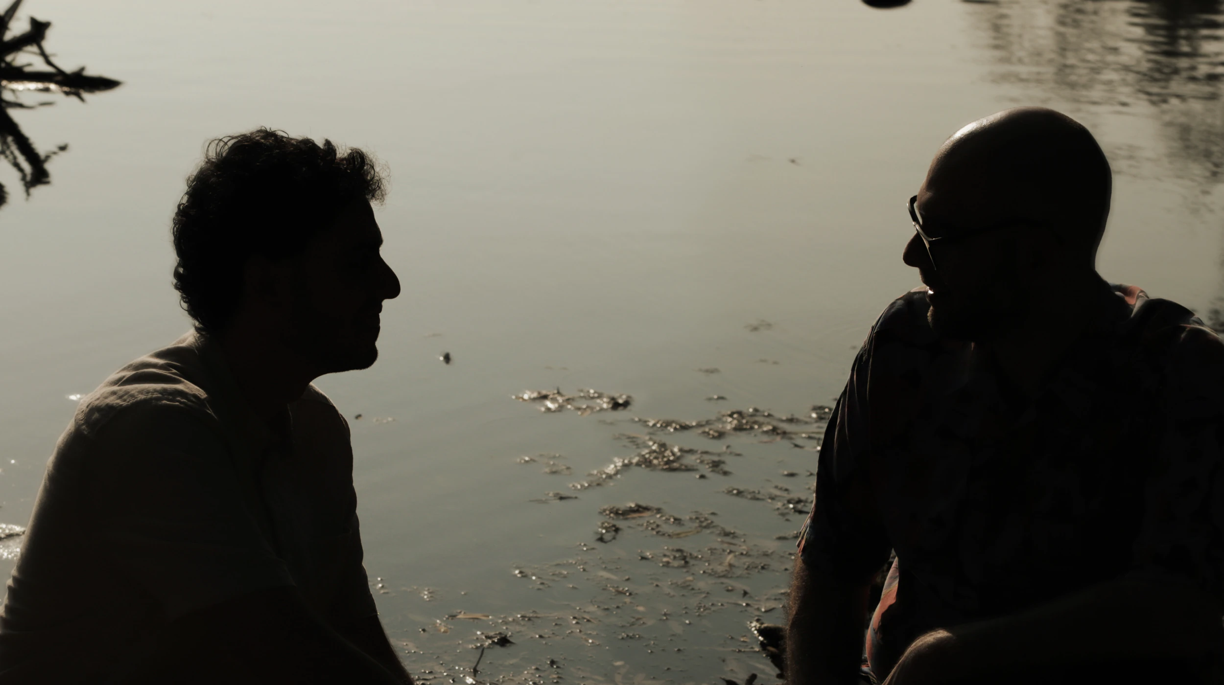 Silhouettes of Antonis and Savvas sitting by the water at sunset, talking quietly. Photo by Artemis Evlogimenou