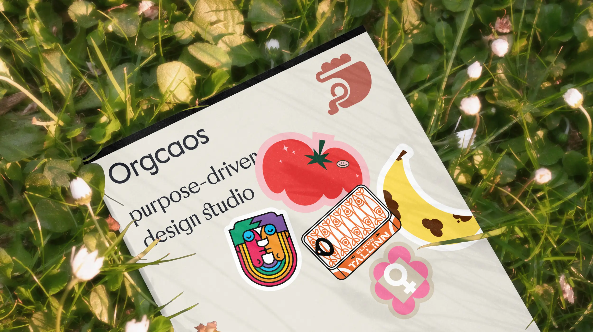 Sticker-covered Orgcaos brand card lying in grass with playful illustrations of fruit and symbols