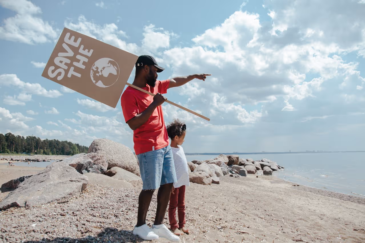 A man holding a sign that says Save the Planet and a child standing on a rocky beach. A man is pointing in the distance. 