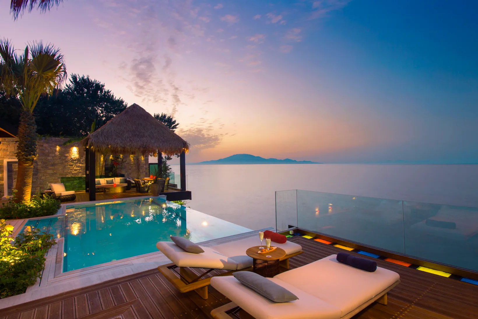 Luxury infinity pool and lounge chairs on wooden deck overlooking calm sea with island silhouette at sunset.