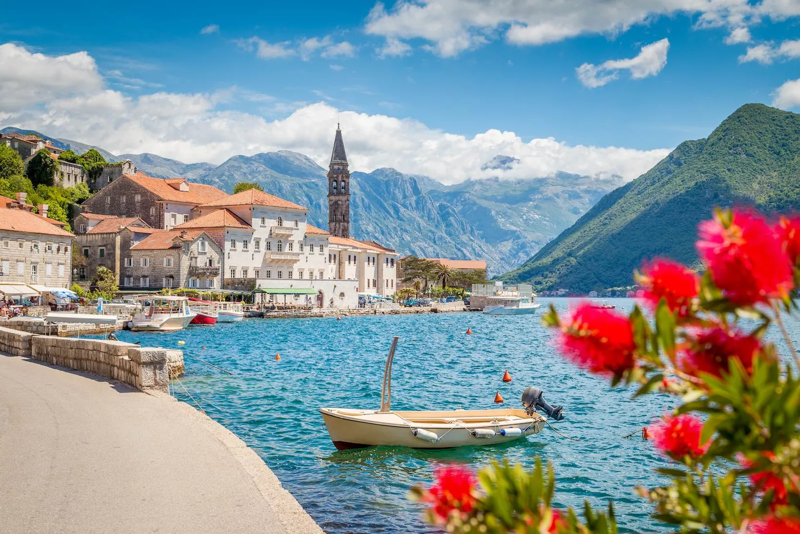 Coastal town with stone buildings and a tall bell tower by blue water, small boat floating, and red flowers in foreground.
