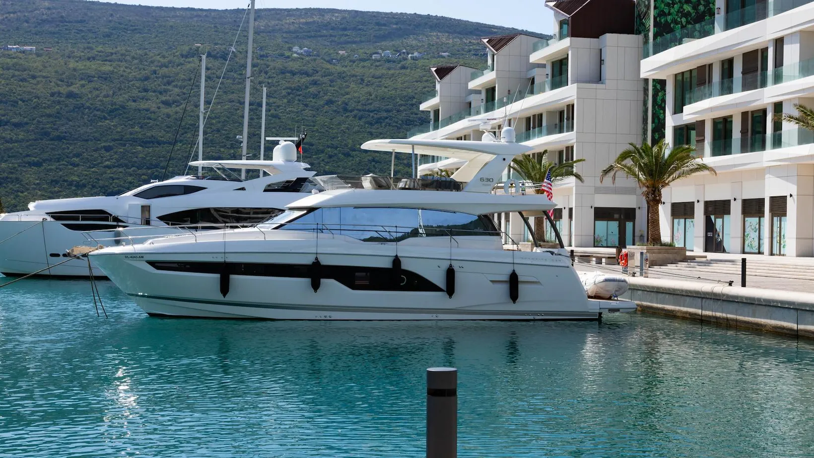 Two white luxury yachts docked in calm turquoise water beside modern buildings with palm trees and a green hillside in the background.