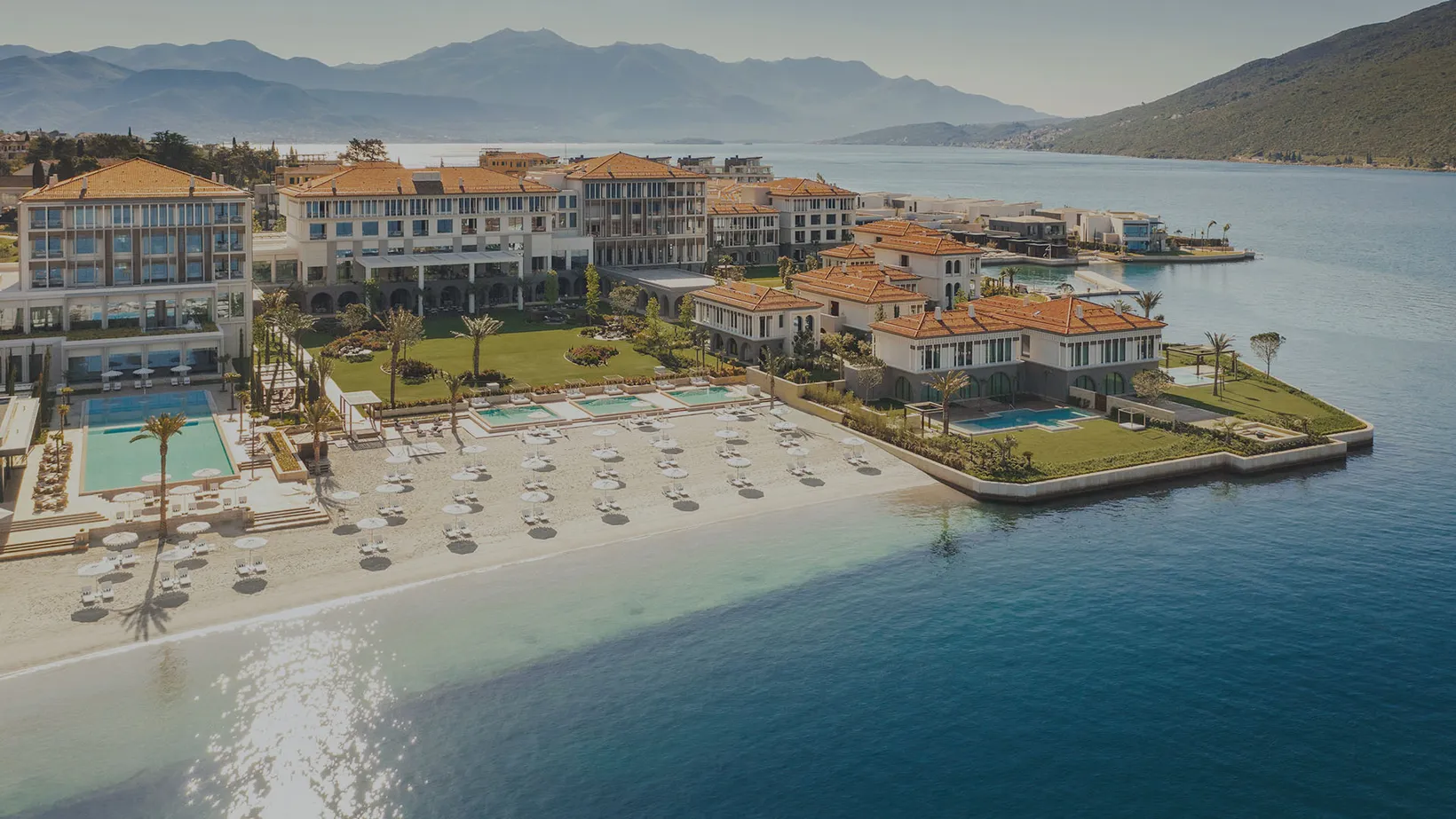 Luxury beachfront resort with multiple pools, sandy beach with umbrellas, and mountains in the background.