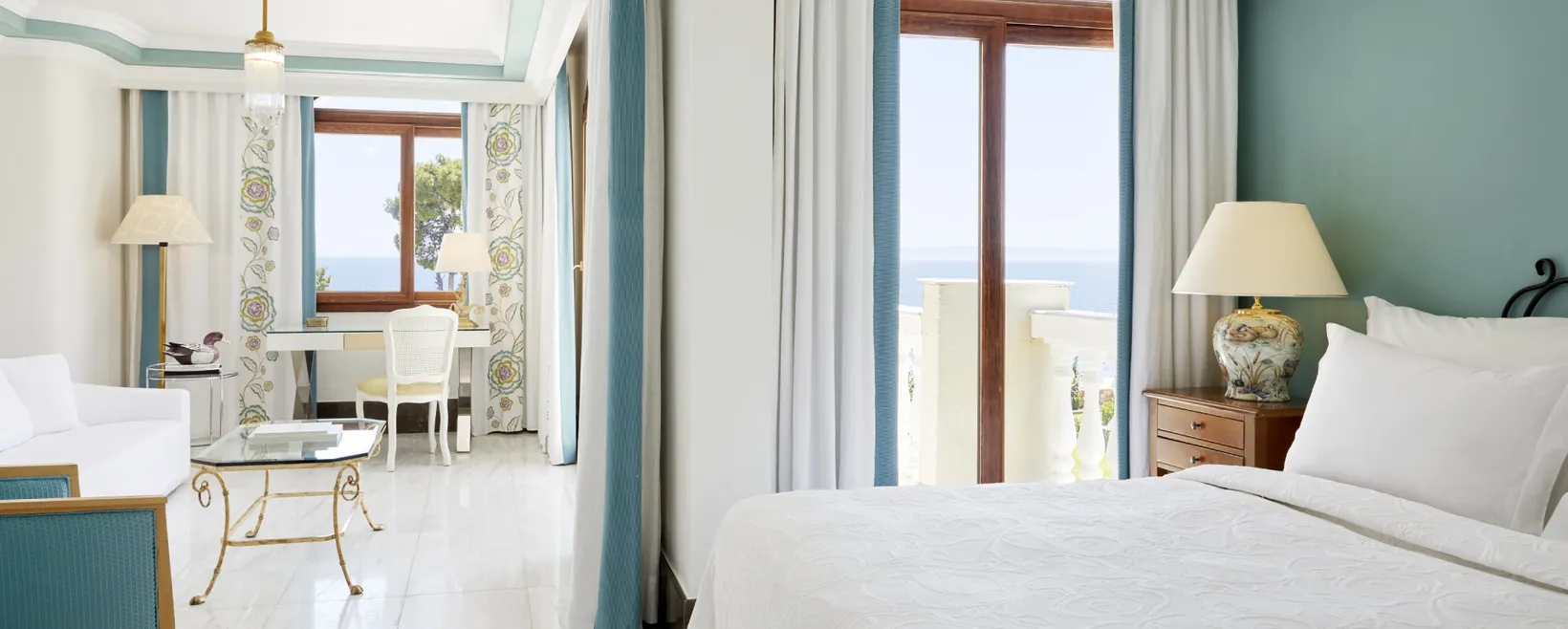 Bright hotel suite with a white bed, wooden nightstand with lamp, seating area, desk, and window showing ocean view.