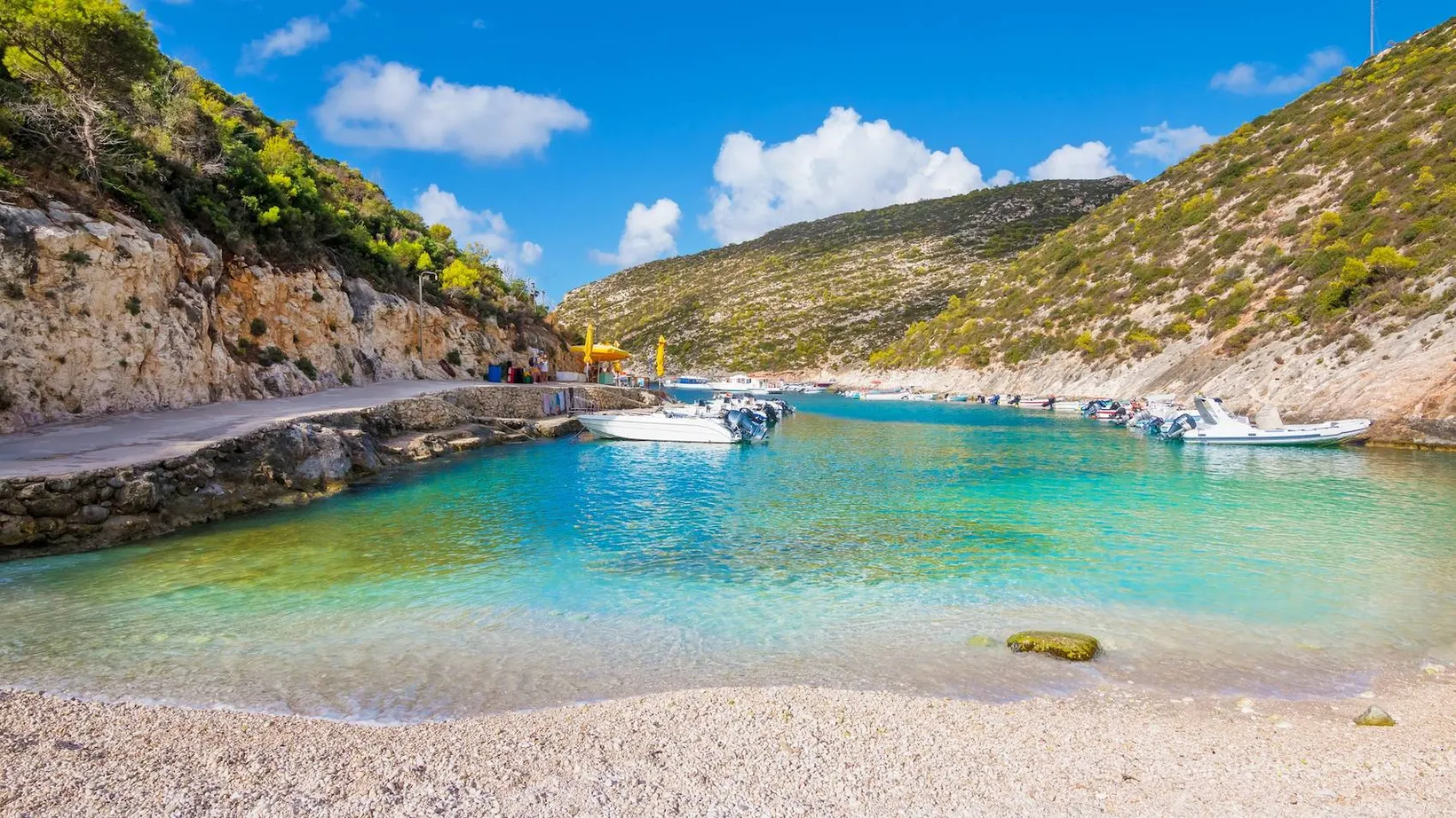 Clear turquoise water bay surrounded by rocky hills with small boats docked along a stone pier under a partly cloudy blue sky.