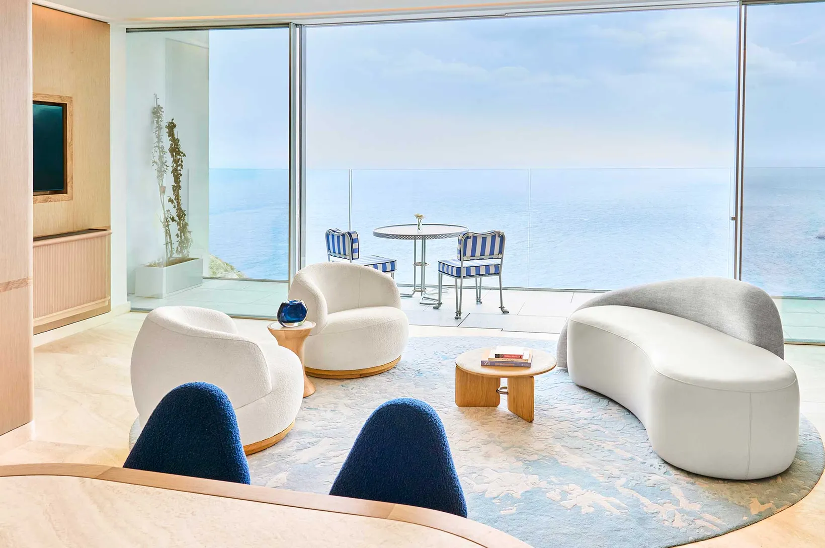 Modern living room with white curved chairs and sofa, small wooden tables, and floor-to-ceiling windows overlooking a balcony and ocean view.