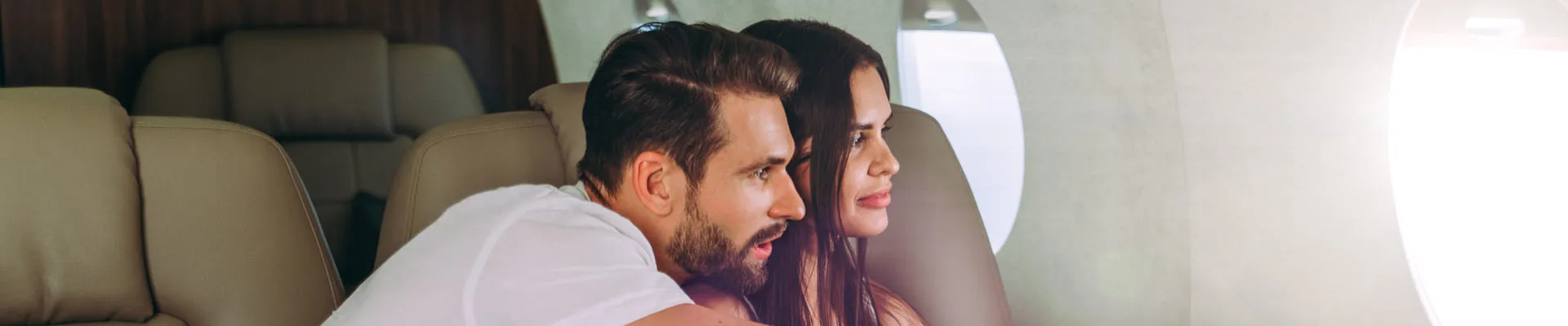 Couple sitting close together and looking out the window inside a private jet with beige leather seats.