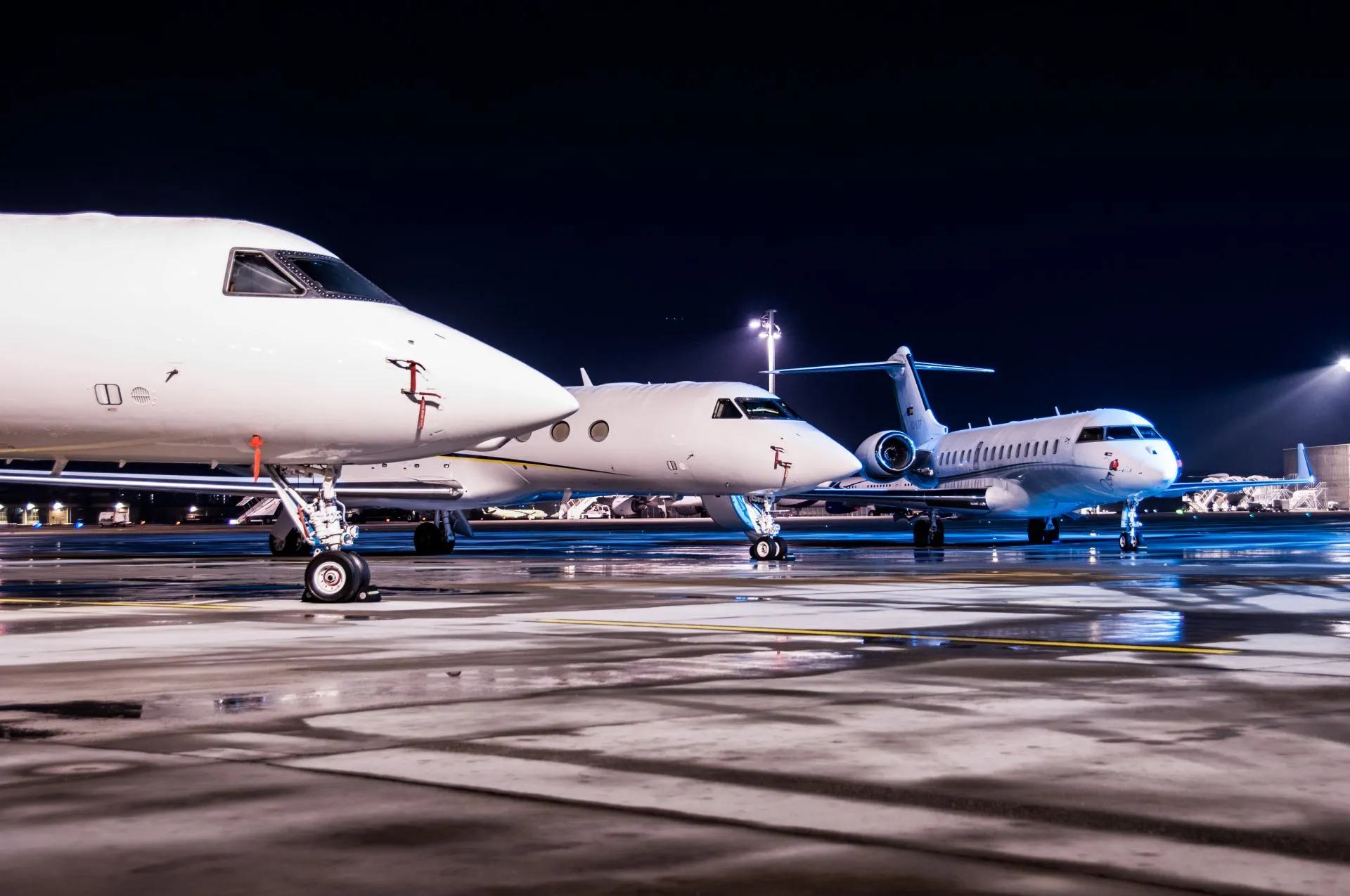 Three white private jets parked on a wet tarmac at night under bright airport lights.