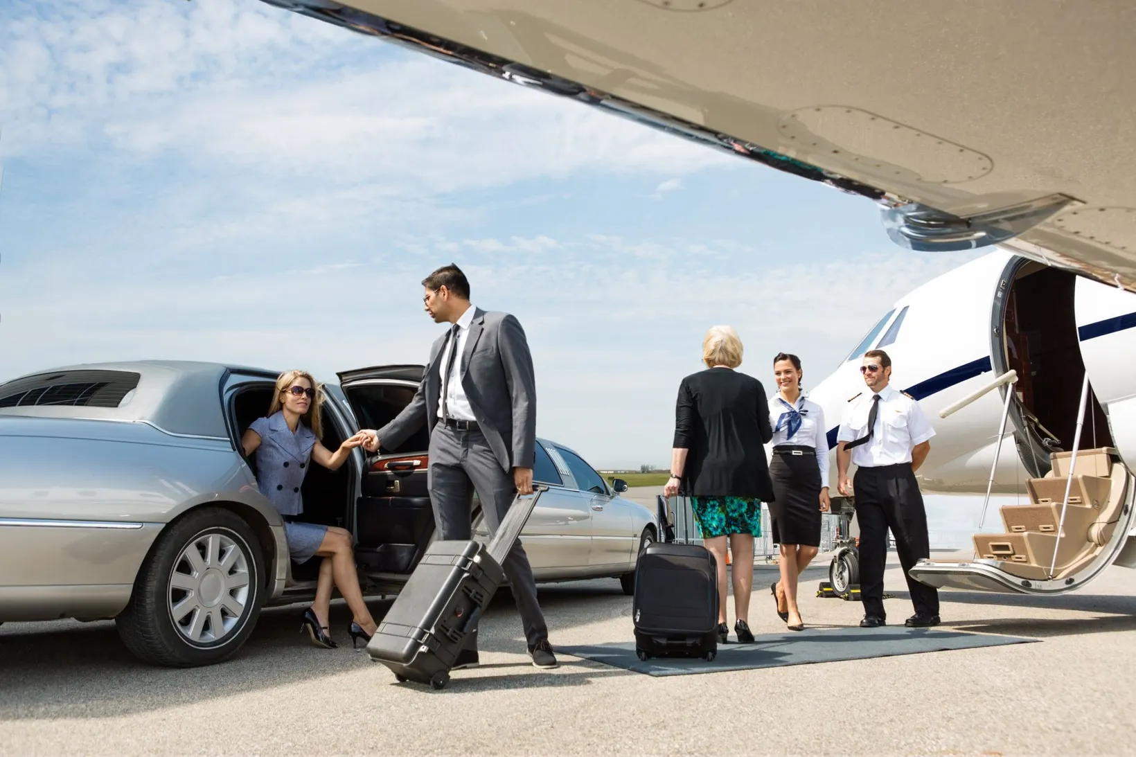 Businesswoman getting out of a limousine while a man in a suit helps her, with a woman pulling luggage toward a private jet where a female flight attendant and male pilot stand by the entrance.
