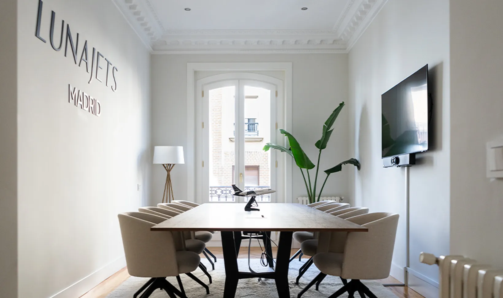 Modern conference room with a long wooden table, beige swivel chairs, a model airplane centerpiece, a floor lamp, large potted plant, and wall-mounted TV.