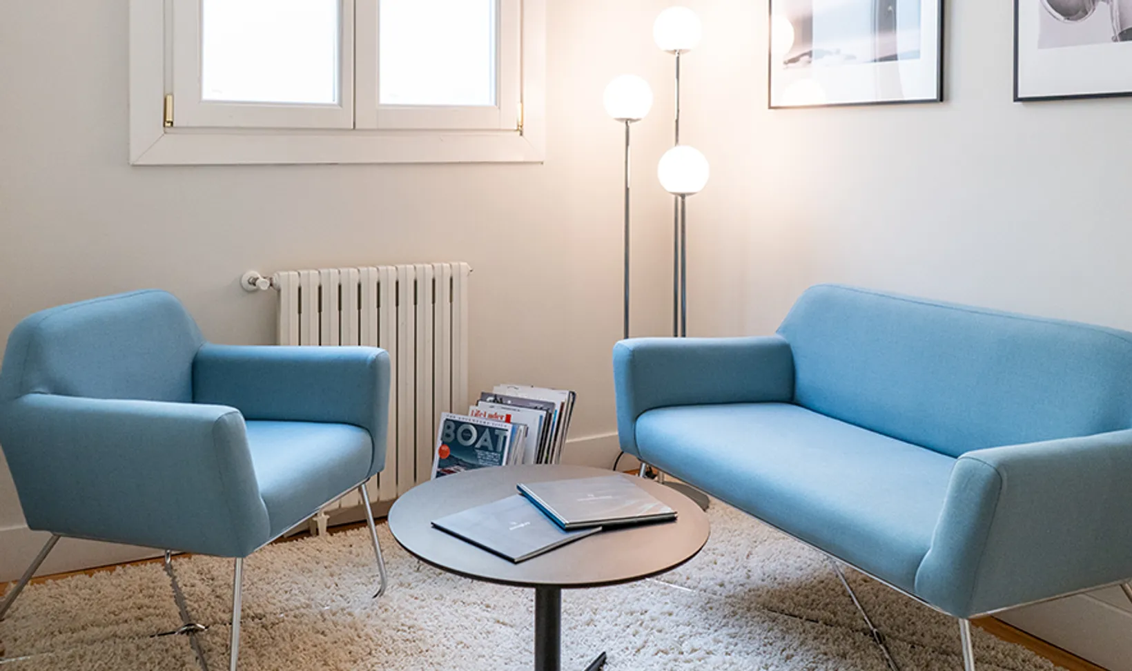 Modern office seating area with a light blue armchair, matching sofa, round table with books, and floor lamp near a window.