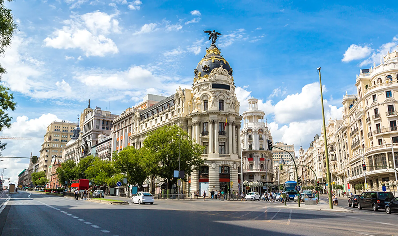 Busy urban street in Madrid with historic buildings, green trees, pedestrians, cars, and a bright blue sky with clouds.