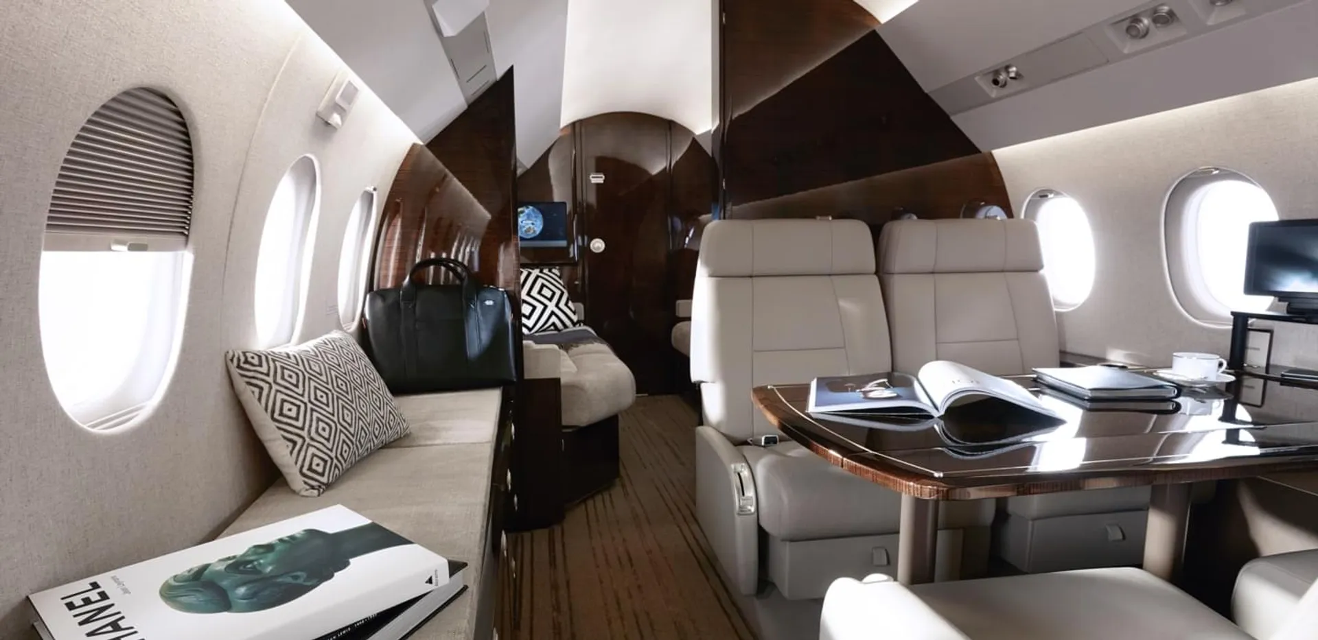Luksusowe wnętrze prywatnego odrzutowca z beżowymi skórzanymi siedzeniami, drewnianym stołem, sofą z wzorzystymi poduszkami i oknami wzdłuż kabiny.